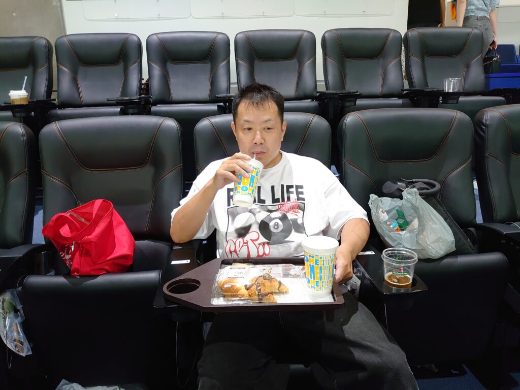 映画館のシートでフードトレイを使いながら食事を楽しむ男性。ポップコーンやドリンクが並び、くつろいだ雰囲気の中で映画鑑賞前のひととき