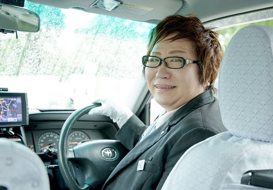 タクシー車内で笑顔を見せる女性ドライバー。運転席に座り、安全運転と親しみやすさを感じさせる雰囲気。