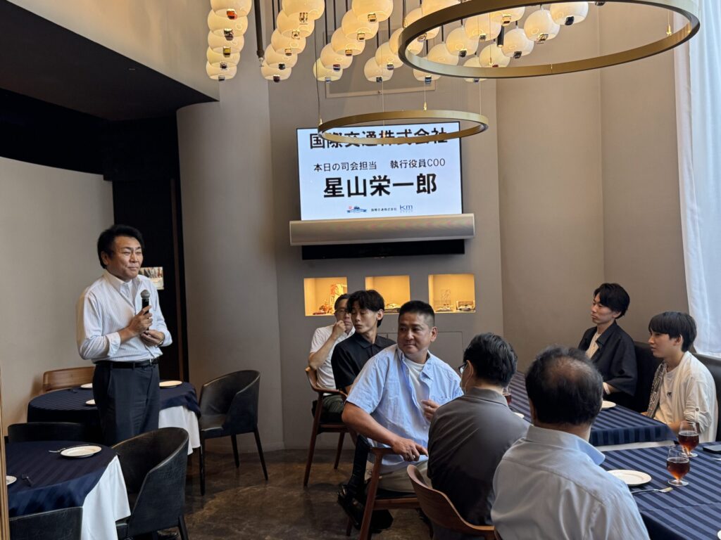 国際交通株式会社の懇親会で挨拶する執行役員COOの星山栄一郎氏。会場には社員が集まり、和やかな雰囲気で話を聞いている場面。
