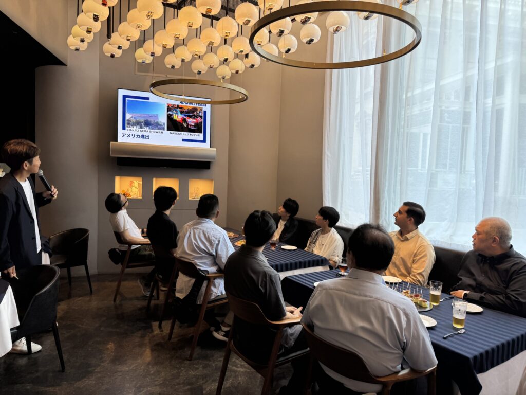 国際交通株式会社の懇親会でプレゼンテーションを聞く参加者たち。スクリーンに映し出された資料を見ながら真剣に耳を傾けている様子。
