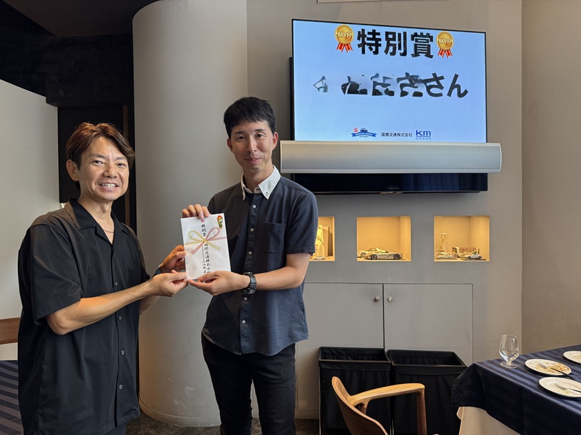 国際交通株式会社の社内イベントにて、特別賞を受賞した社員が表彰者と並び賞状を受け取っている場面の写真